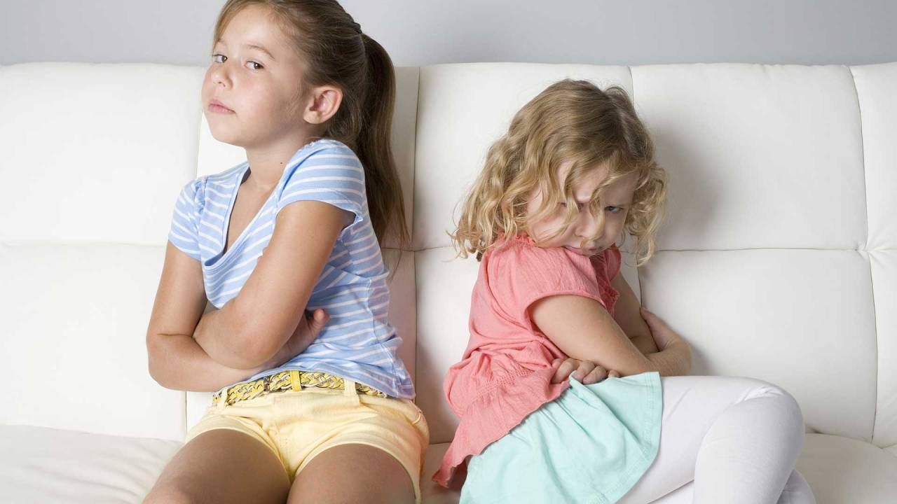 duas meninas, sentadas em um sofá, uma de costas para a outras, com expressões de brava