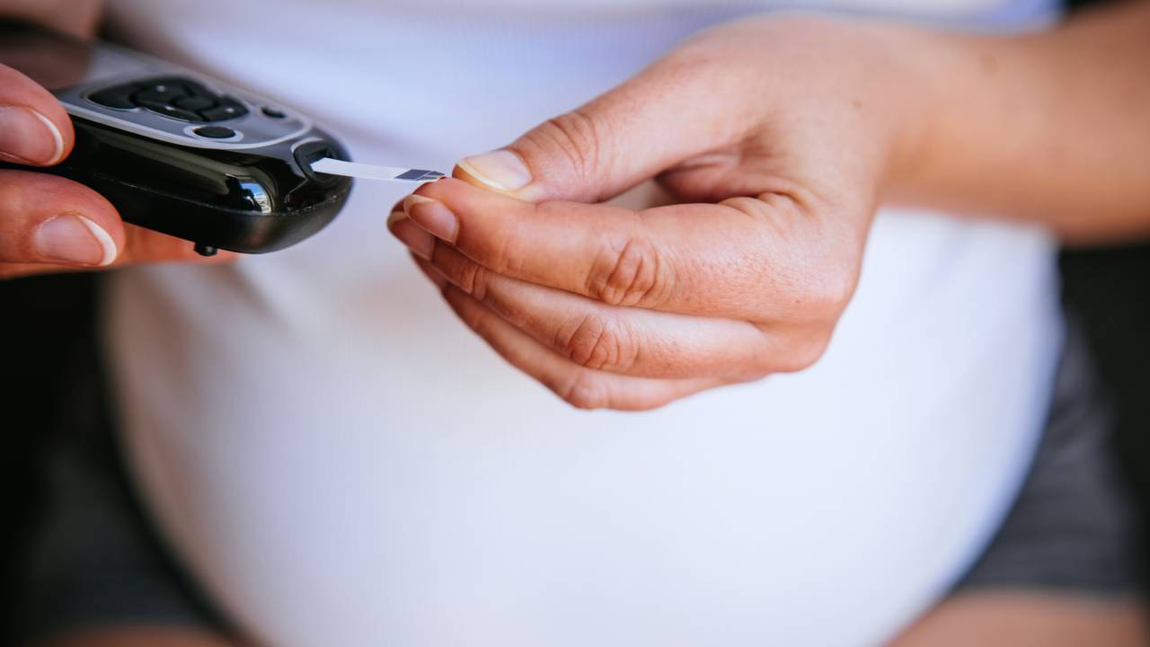 Na imagem, parte do corpo de uma mulher grávida. Aparece a barriga, usando camiseta branca. Ela está medindo a insulina: segura um aparelho com a mão direita e mede com a mão esquerda. Ela tem a pele branca.