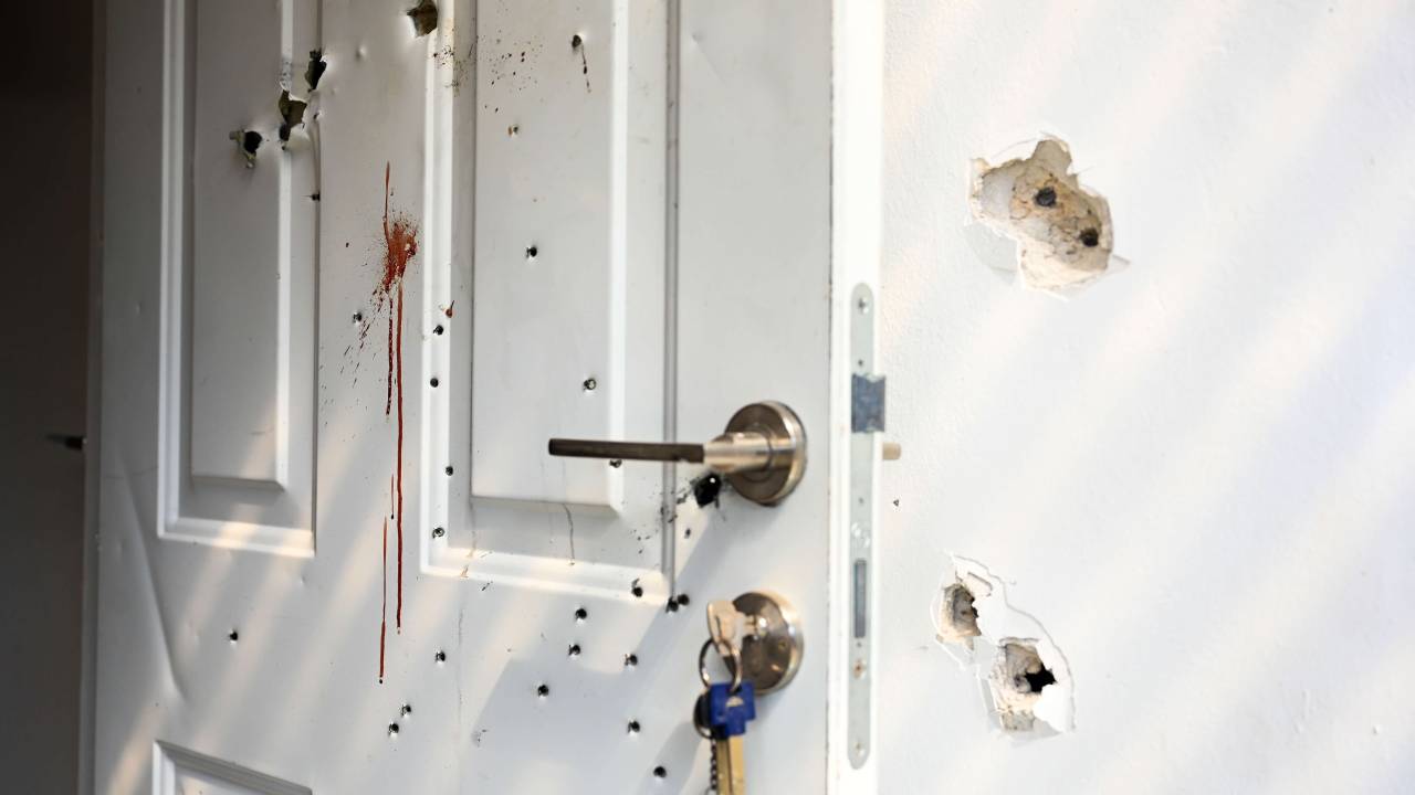 Na foto, uma porta branca, aberta, com a chave na fechadura, repleta de furos de bala. No meio da porta, há uma mancha de sangue escorrida. Na parede branca, ao lado, também há furos de bala.