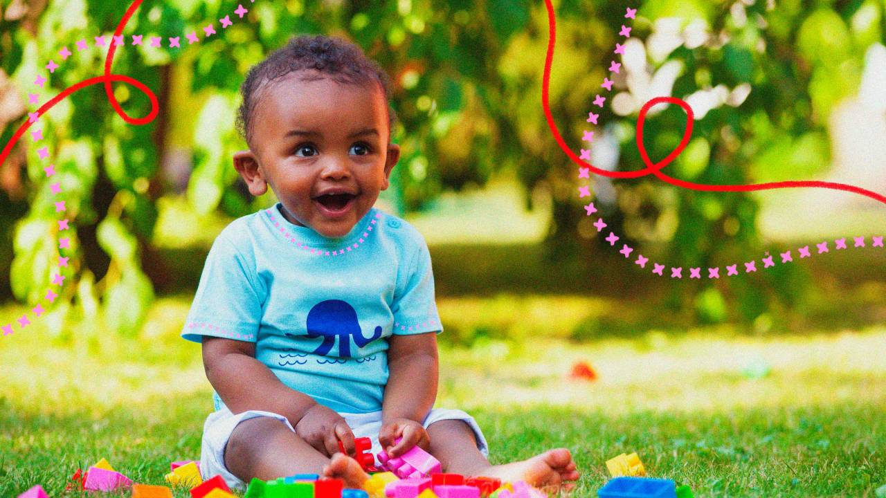 Bebê menino, de pele negra, por volta de 1 ano e meio, veste camiseta azul e shorts branco. Ele está em um jardim, sentado na grama, sorrindo. No chão, à sua frente, há blocos coloridos de montar. No plano de fundo, há árvores. Ele está sorrindo.