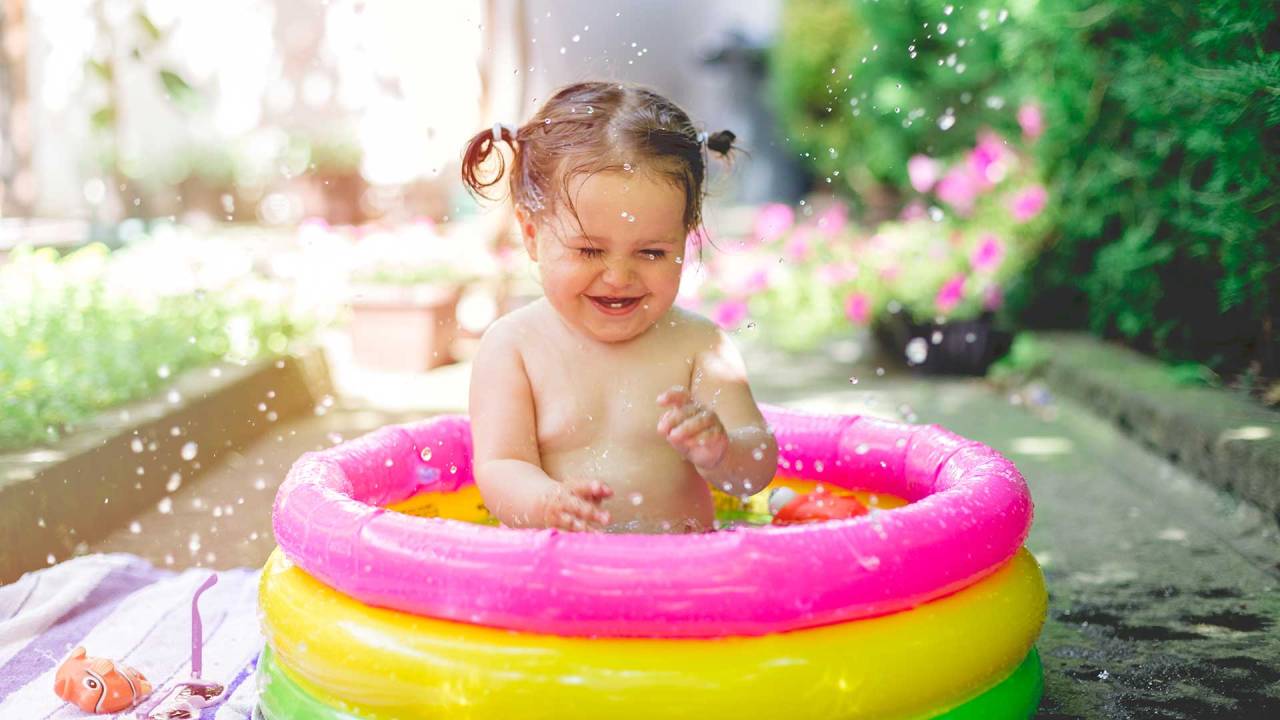 garotinha sentada em uma piscina inflável infantil e colorida. Ela sorri diante dos respingos de água