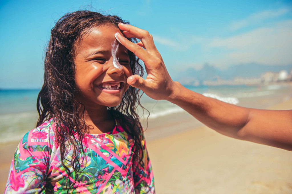 Na foto, uma menina negra na praia, com o mar e o céu de fundo. Ela tem cabelo comprido e cacheado, está sorrindo e vestindo um maiô de manga comprida e bem colorido. A mão de uma pessoa adulta passa protetor solar, um produto branco, no rosto dela.