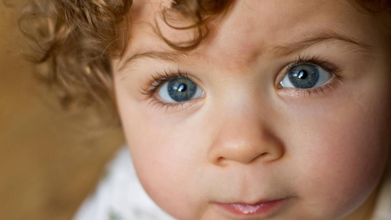 Bebê, por volta de 1 ano e meio, pele clara, olhos bem azuis, cabelo encaracolada castanho claro ou loiro escuro. A foto está bem fechada no rostinho dele, que olha para a foto e está sério.