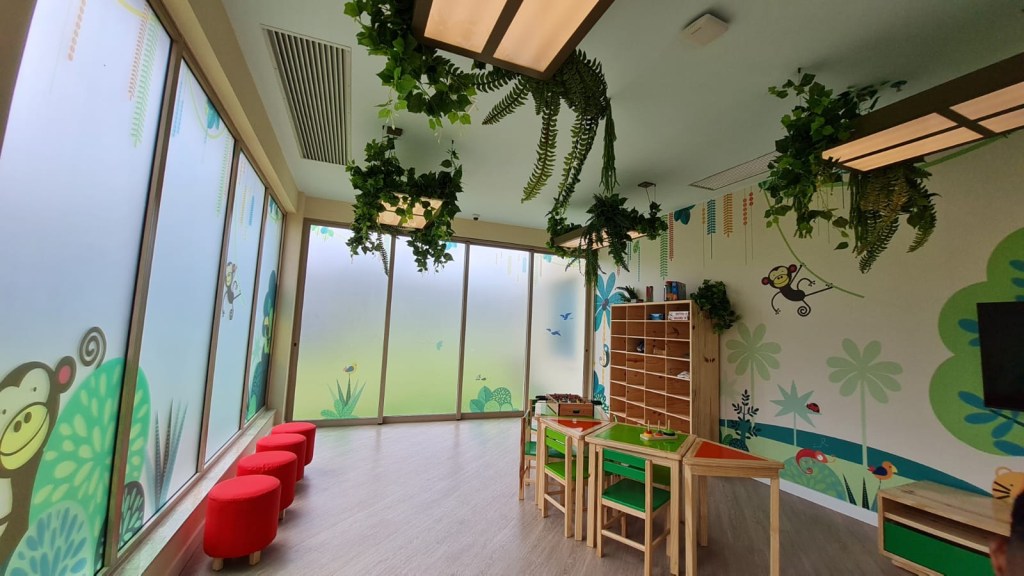 Área interna do Star Camp, com mesas coloridas de plastimadeira, bancos vermelhos, chão claro, adesivos de macacos e plantas nas paredes e plantas penduradas na luminária.