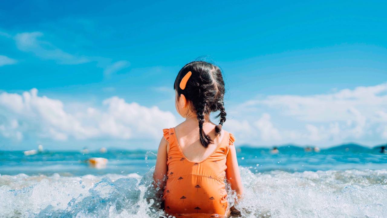 Na foto, uma menina de costas, sentada na beira do mar, com a onda cobrindo suas pernas. Tem a pele clara, cabelo castanho escuro comprido preso para trás em duas tranças. Usa uma presilha laranja do lado direito da cabeça e um maiô laranja. O céu está bem azul.