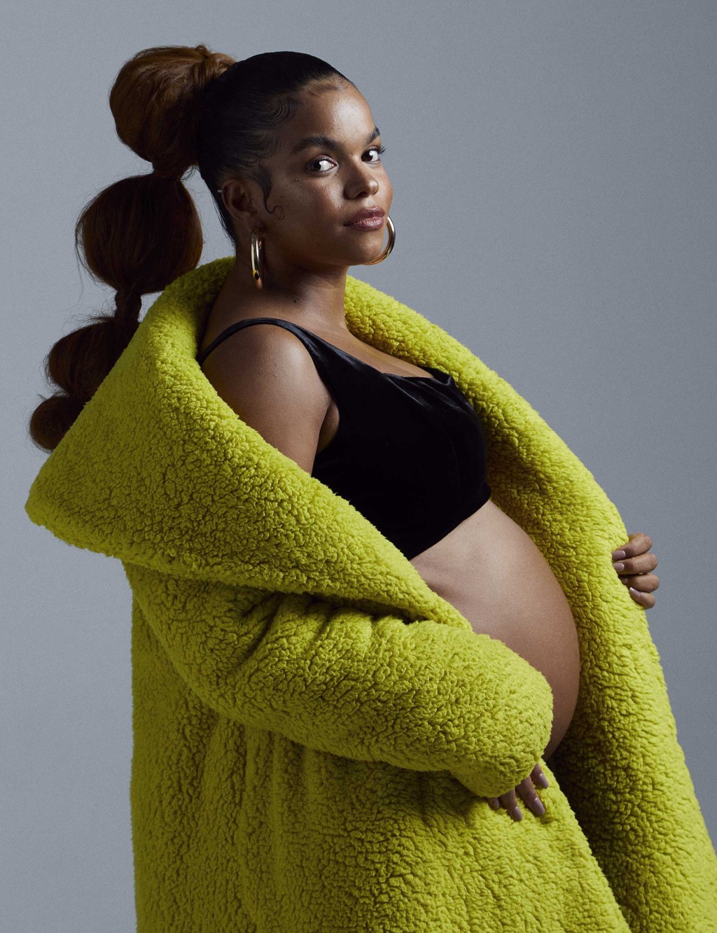 mulher grávida olhando com expressão séria para a câmera. Ela usa um casaco aberto e um top, deixando a barriga à mostra