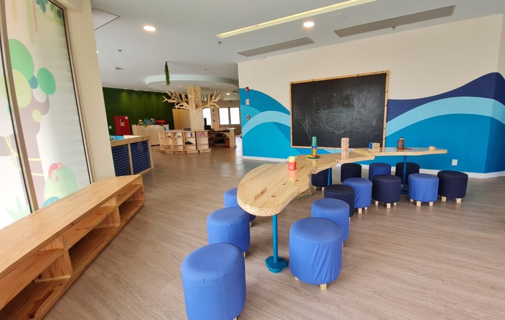 Na foto, uma área interna com mesa, puffs azuis e redondos, uma mesa clara e copmrida, lousa na parede, chão claro e estante de madeira.