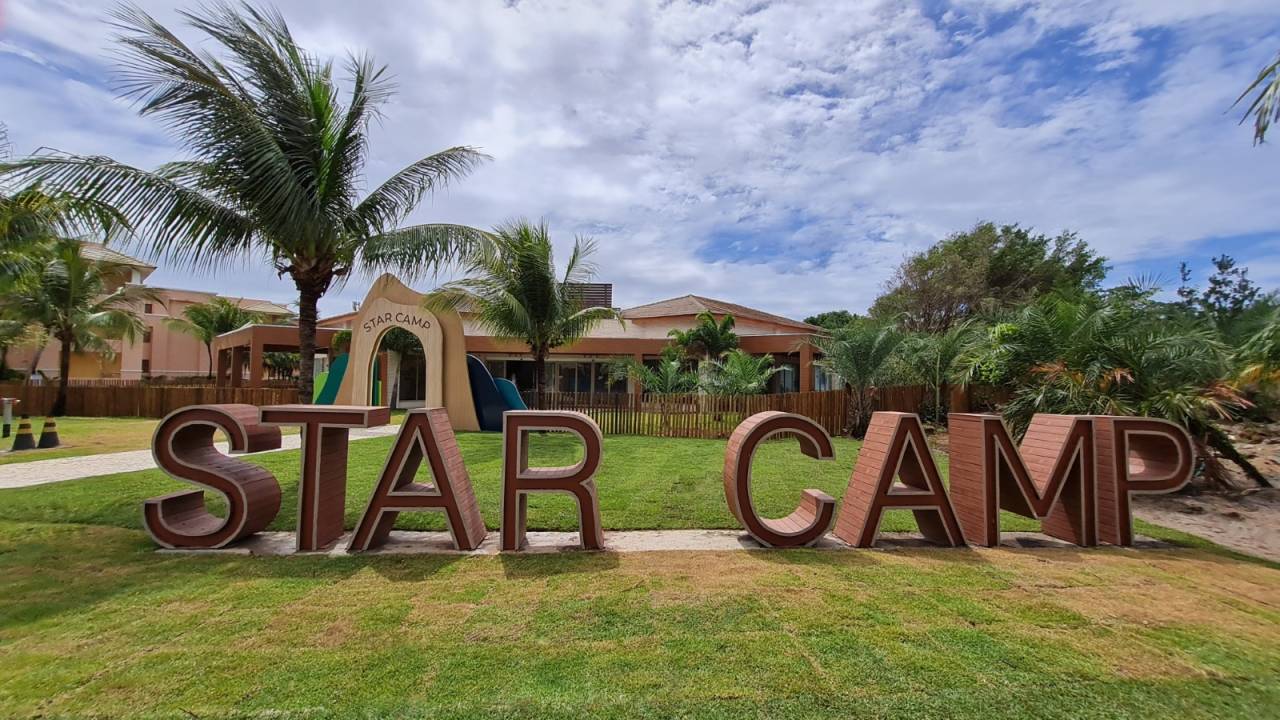 Na imagem, um letreiro em cor de madeiro escrito STAR CAMP. Está em um gramado, com coqueiros. Atrás, é possível ver uma construção baixa, mais árvores e um céu nublado.