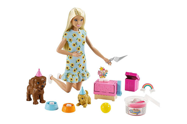 Boneca Barbie ajoelhada, usando um vestido floral. Perto dela estão cachorrinhos de brinquedo e outros objetos coloridos.