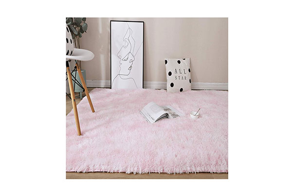 Chão de madeira onde há um tapete rosado felpudo. Sobre eles estão um livro, um quadro, uma xícara e uma almofada