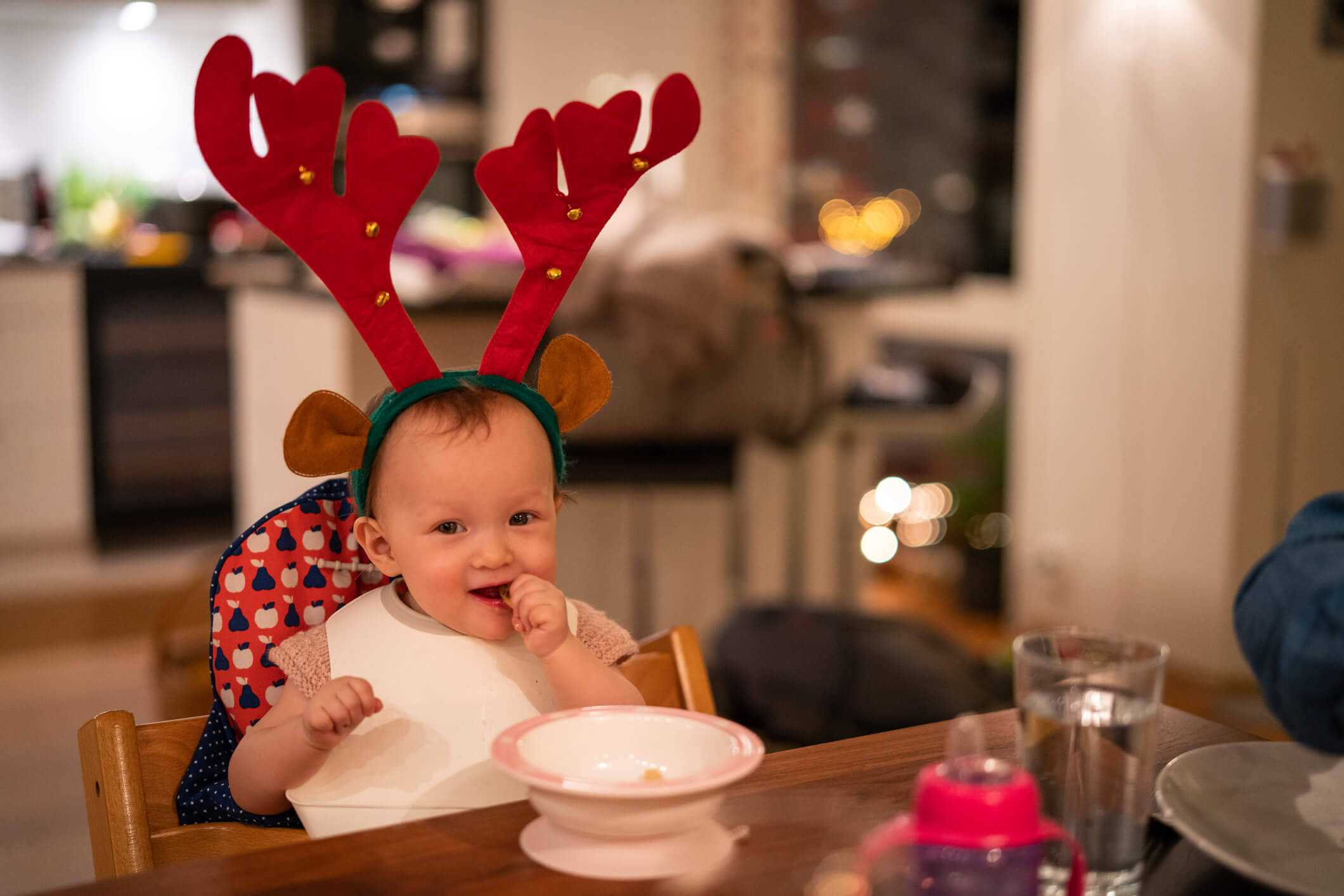 Bebê sentado à mesa, em frente a um pratinho de comida. Ele tem a pele clara, está sorrindo e colocando algum alimento na boca e usa duas grandes orelhas de renas vermelhas.