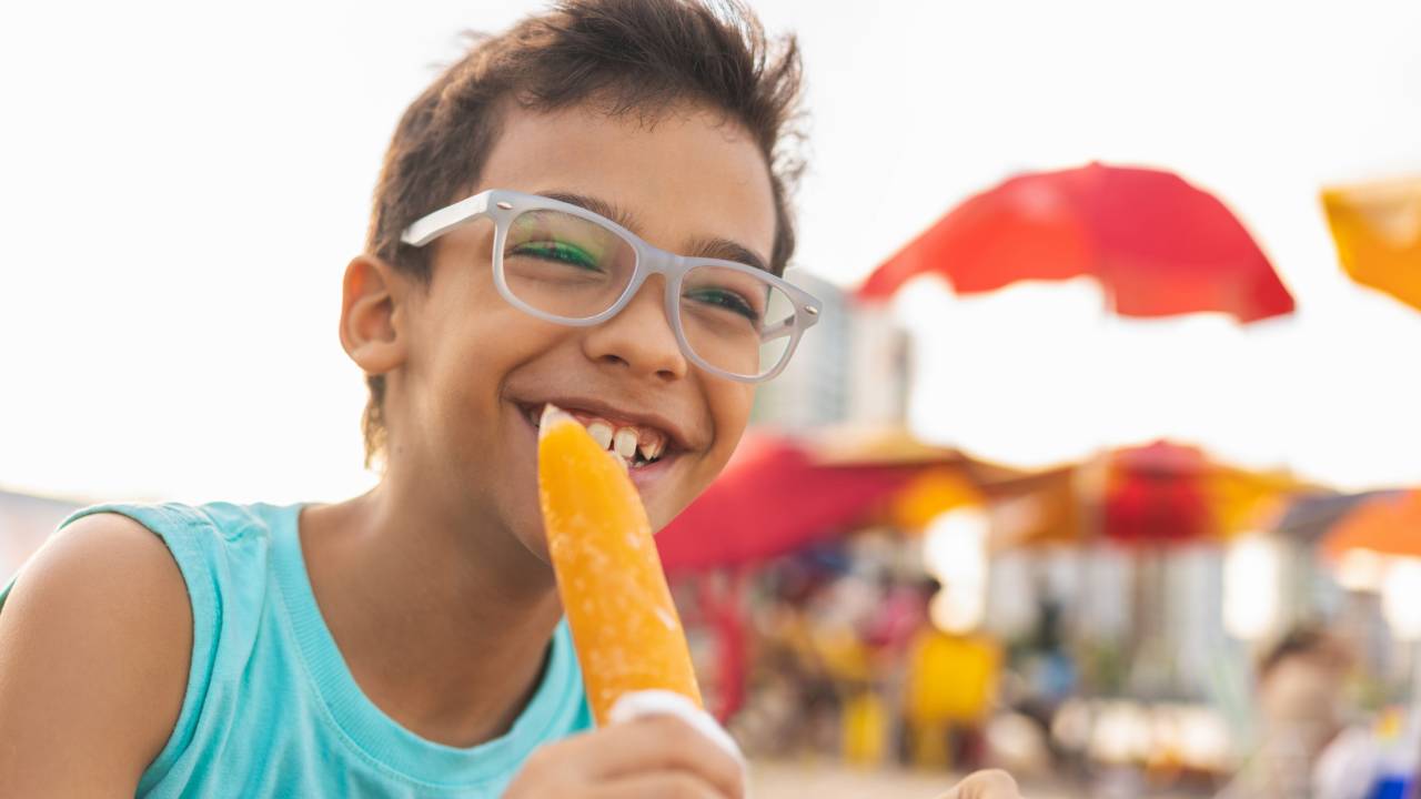 Foto de um menino, por volta de 10 anos, na praia, chupando geladinho cor de laranja. Ele em o cabelo curto, castanho e usa óculos. Usa camiseta azul turquesa.