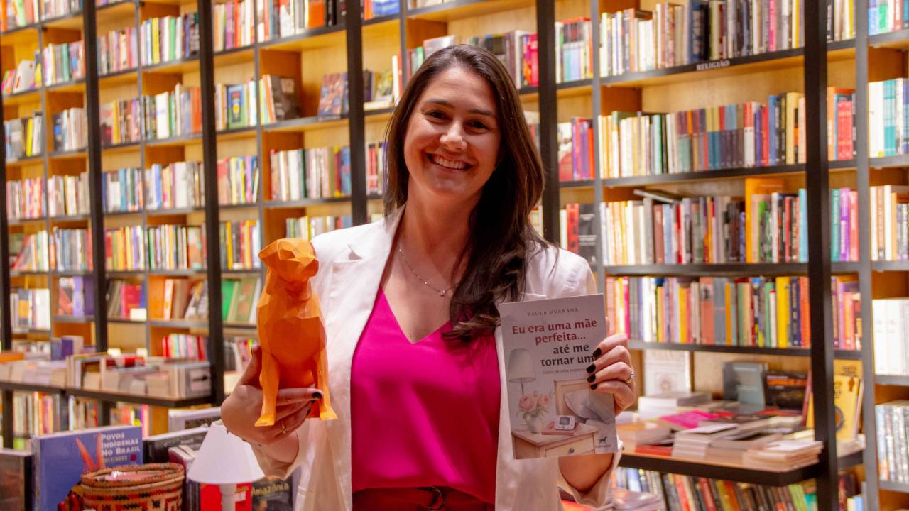 Paola Guaraná é uma mulher branca, de cabelos castanhos, lisos e compridos. Está em uma livraria, com livros ao fundo. Segura um livro e uma escultura de cachorro. Veste roupa rosa.