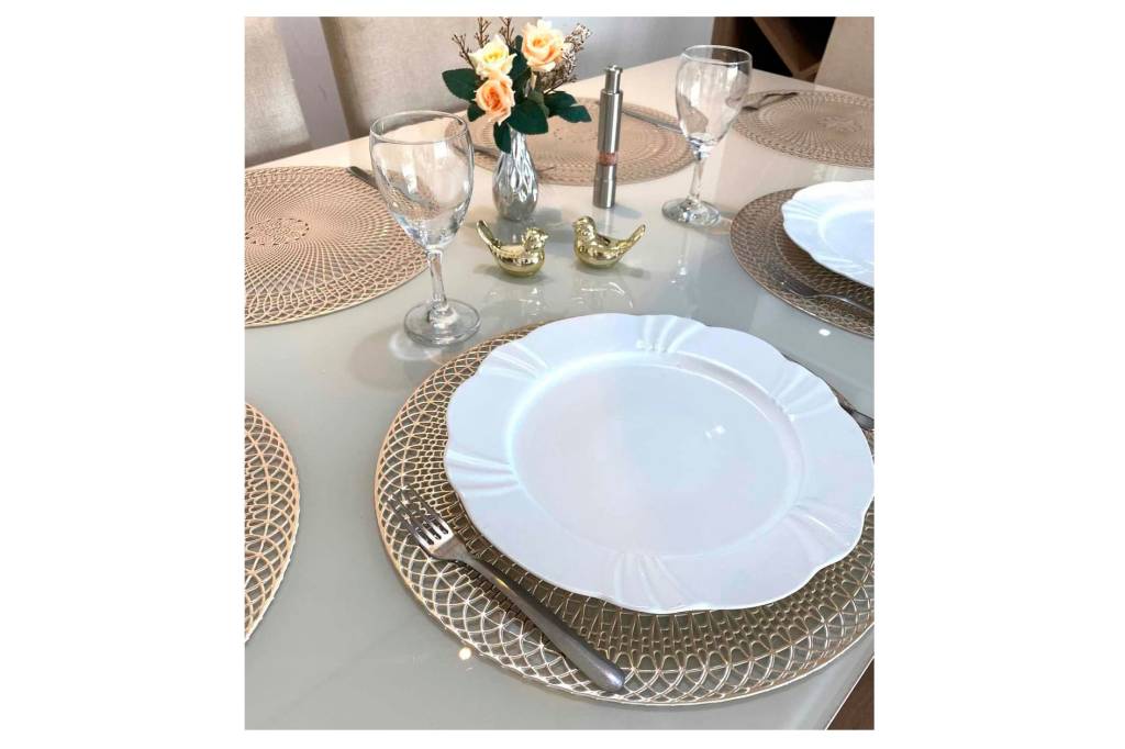 Sobre fundo branco, foto de uma mesa posta. Tem suportes redondos e dourados para pratos brancos. Taças de vidro e talheres prateados.