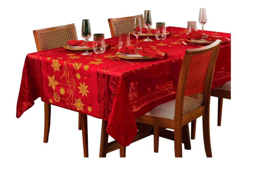 Toalha vermelha com detalhes dourados sobre mesa de madeira retangular. Há cadeiras em volta e o fundo é branco.