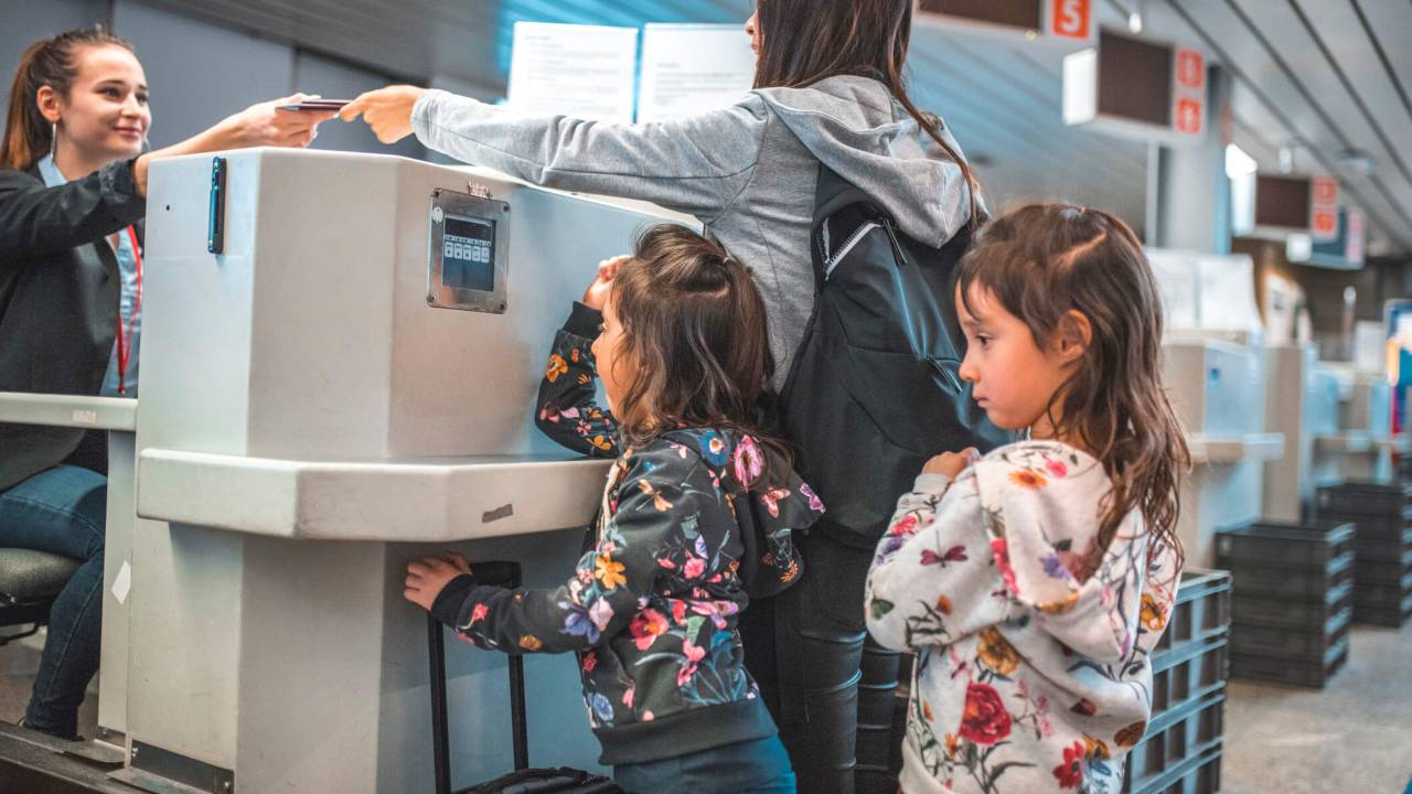 Mãe e filhas em balcão de check-in de aeroporto, entregando passaportes à atendente.