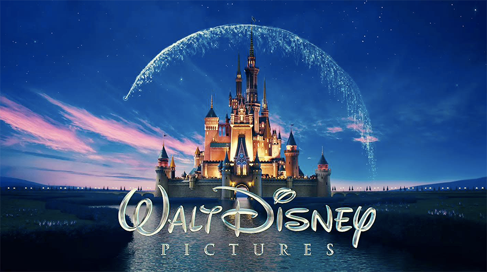 -Logo da Disney - um castelo no centro da imagem, iluminado, com o rastro de uma estrela semicircular sobre ele. No fundo, um céu azul e, à frente, um lago.